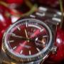 Eine Rolex Day-Date mit tiefrotem Zifferblatt, umgeben von glänzenden Kirschen. Das kräftige Rot des Hintergrunds harmoniert perfekt mit dem Sonnenschliff-Zifferblatt und hebt die Uhr als kraftvolles Statement-Piece hervor.