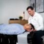 Ein Chiropraktiker führt eine manuelle Therapie an einem Patienten durch, der auf einer Behandlungsliege liegt. Der Chiropraktiker, in einem weißen Hoodie gekleidet, hält sanft den Kopf des Patienten, um eine Anpassung an der Halswirbelsäule vorzunehmen. Der Patient trägt ein hellblaues Hemd und liegt entspannt mit geschlossenen Augen auf dem Rücken. Im Hintergrund befinden sich ein Holzmöbelstück mit einer Pflanze und einer Lampe sowie medizinische Poster, die die Anatomie des menschlichen Rückens und des Nervensystems darstellen. Ein Chiropraktiker führt eine manuelle Therapie an einem Patienten durch, der auf einer Behandlungsliege liegt. Der Chiropraktiker, in einem weißen Hoodie gekleidet, hält sanft den Kopf des Patienten, um eine Anpassung an der Halswirbelsäule vorzunehmen. Der Patient trägt ein hellblaues Hemd und liegt entspannt mit geschlossenen Augen auf dem Rücken. Im Hintergrund befinden sich ein Holzmöbelstück mit einer Pflanze und einer Lampe sowie medizinische Poster, die die Anatomie des menschlichen Rückens und des Nervensystems darstellen.