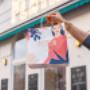 PLAIN Vienna To-go: Hand mit gebrandeter PLAIN-Papiertragetasche vor dem Café mit Neonlogo und Schanigarten in 1090 Alsergrund. Ideal für Take-away, Brunch & Coffee to go – stylisches Branding und urbanes Flair in Wien.