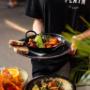 Servicemoment im PLAIN Vienna: schwarze Logo-Shirt-Crew serviert eine gusseiserne Pfanne mit Garnelen in Tomatensauce, Avocado & Kräutern, Brotscheiben am Rand; vorne Bowl mit Couscous & Zitrone, Aperitivo daneben. Outdoor-Service, 1090 Wien.