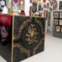 Der Empfangstresen mit kunstvoller, in Gold und Schwarz gestalteter Frontverzierung steht im Mittelpunkt. Dahinter sind zahlreiche Wandbilder, Banner und kunstvolle Details angebracht, die sofort die kreative und extravagante Stimmung des Studios widerspi